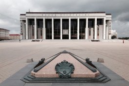 Duży biało-kremowy gmach z wysokimi kolumnami i przeszklonym wejściem stoi na szerokim pustym placu pod pochmurnym niebem, a na pierwszym planie widać trójkątny granitowy pomnik z metalową tarczą i dwiema kotwicami.