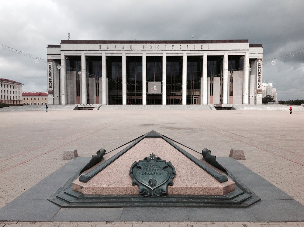 Monumentalny budynek z wysokimi kolumnami stojący na rozległym, pustym placu; na pierwszym planie kamienny trójkątny pomnik z ozdobną metalową tablicą i dwoma krótkimi działkami, w tle pochmurne niebo i nieliczni przechodnie.