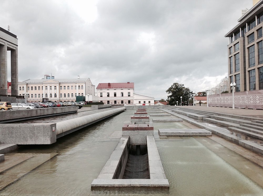 Plac miejski z długimi prostokątnymi kaskadowymi basenami z wodą i betonowymi pomostami na pierwszym planie, po lewej zaparkowane samochody i klasyczne kamienice, po prawej nowoczesny budynek biurowy i schody, w tle drzewa oraz zachmurzone niebo.