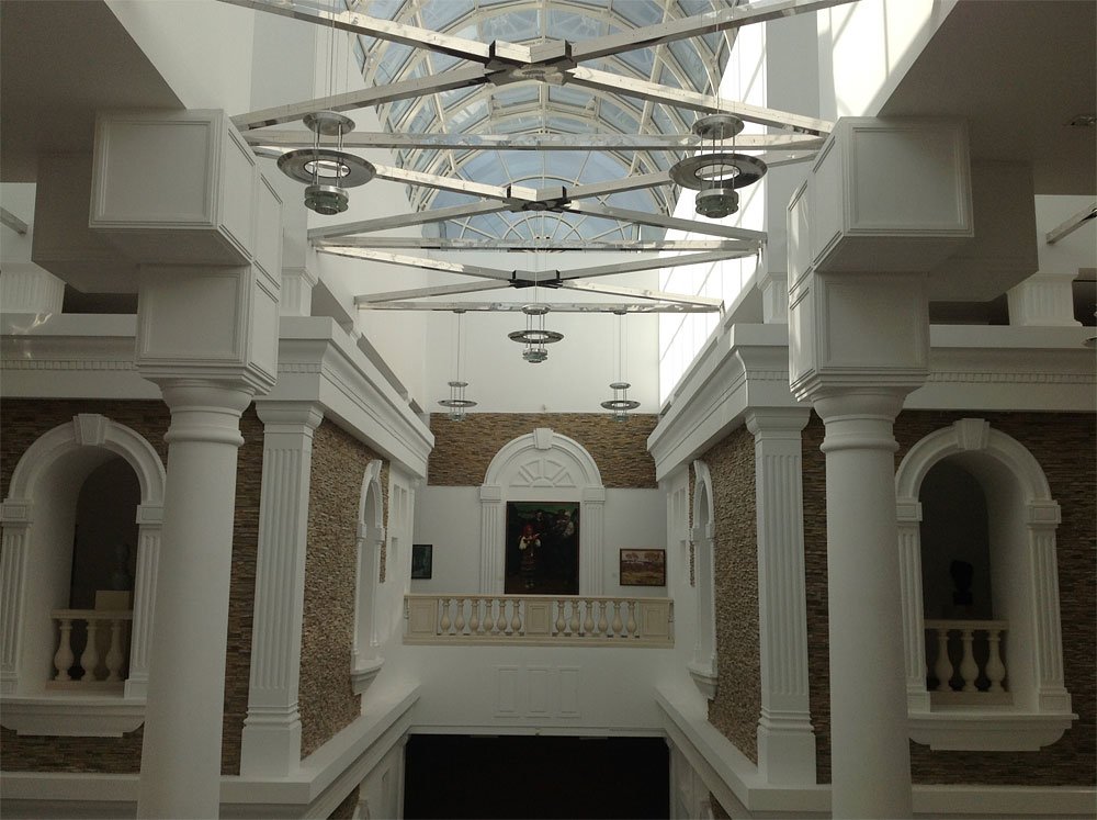 Wnętrze atrium z białymi klasycznymi kolumnami i gzymsem, bocznymi ścianami obłożonymi kamienną okładziną, centralnym balkonem z balustradą i obrazami na tylnej ścianie oraz dużym przeszklonym świetlikiem sufitowym z metalowymi dźwigarami i wiszącymi okrągłymi lampami.