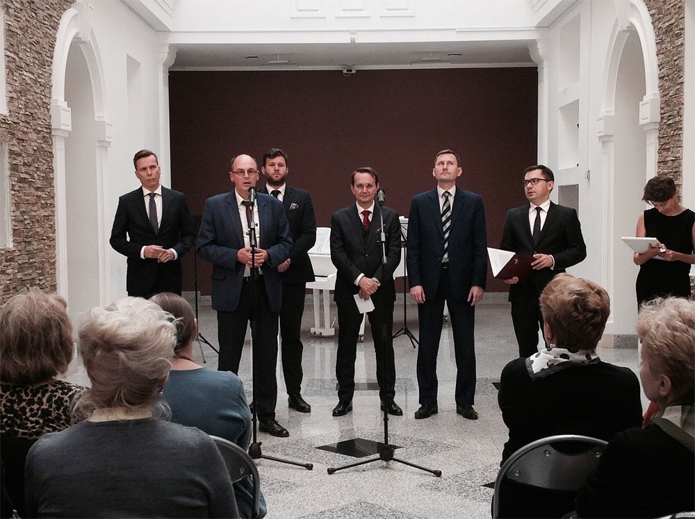 Sześciu mężczyzn w garniturach stoi w równym rzędzie przy mikrofonach na środku jasnej sali z białymi arkadami, kamiennymi ścianami i błyszczącą marmurową podłogą, za nimi widać biały fortepian, z przodu siedzi publiczność tyłem do aparatu, a po prawej stoi kobieta trzymająca tablet.
