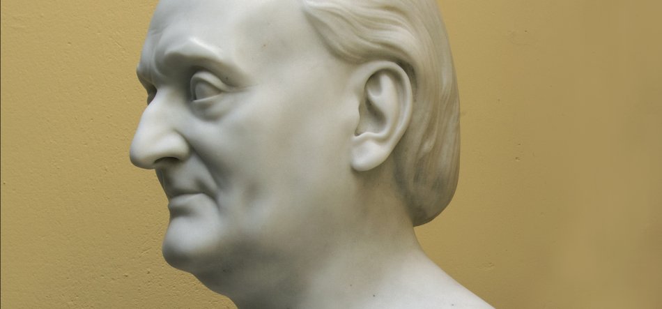 Białe marmurowe popiersie męskiej głowy widoczne z profilu w lewo, z wyraźnym nosem, uchem i zaczesanymi do tyłu włosami oraz widoczną częścią szyi i ramion na jednolitym żółto‑beżowym tle.