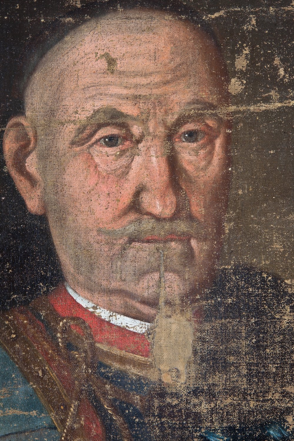 Zbliżenie na fragment starego portretu malarskiego: łysa twarz starszego mężczyzny z niebieskimi oczami, głębokimi zmarszczkami i wąskim, wydłużonym kępkiem brody, w czerwonym kołnierzu i ozdobnym skórzanym pasie na ramieniu; powierzchnia płótna silnie spękana i przetarta, z widocznymi ubytkami farby.