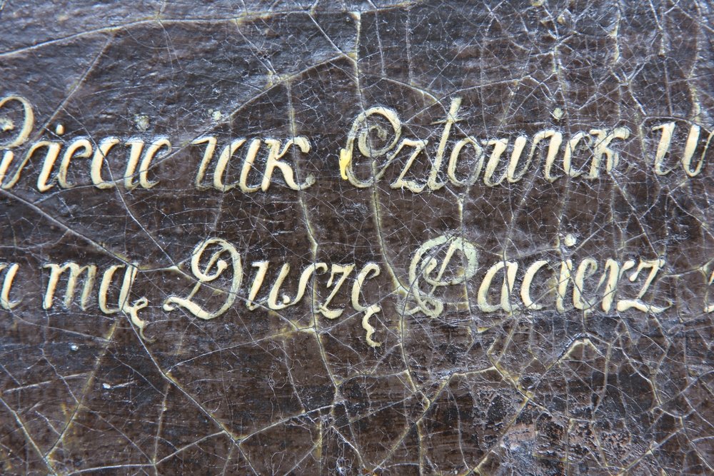 Zbliżenie popękanej ciemnej, lakierowanej powierzchni z ozdobnym złotawym, kaligraficznym napisem w kursywie, na którym widoczne są fragmenty liter i słowo Duszę.