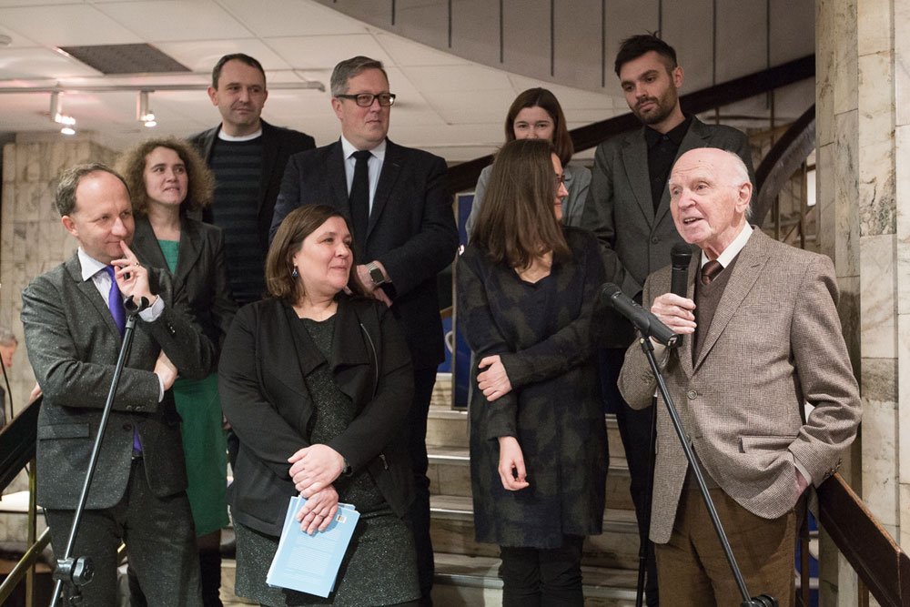 Grupa dorosłych stoi na schodach wewnątrz marmurowego wnętrza; z prawej starszy mężczyzna trzyma mikrofon przy statywie, a inni, ubrani formalnie, stoją obok i za nim, jedna kobieta z przodu trzyma niebieską broszurę.
