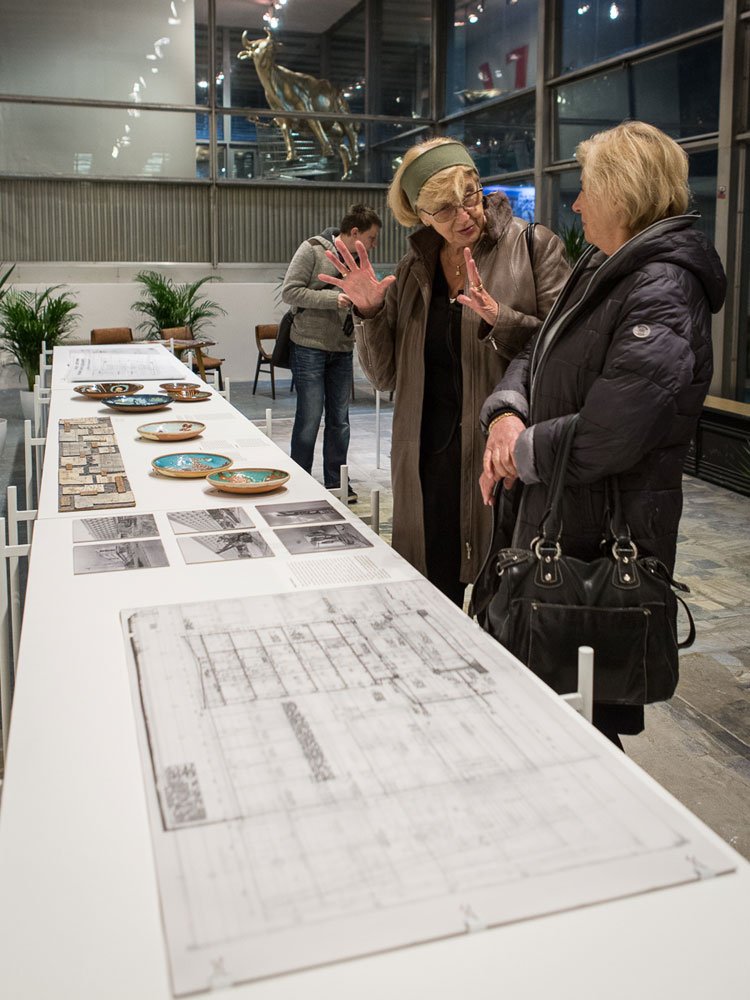 Dwie kobiety w płaszczach rozmawiają przy długim białym stole wystawowym z rozłożonymi planami architektonicznymi, czarno‑białymi zdjęciami, ceramicznymi talerzami i próbką mozaiki; jedna gestykuluje rękami, w tle inna osoba i przeszklona ekspozycja ze złotą rzeźbą zwierzęcia.
