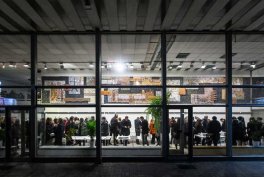 Przez dużą szklaną elewację z metalowymi ramami widać oświetlone wnętrze budynku, gdzie grupa osób stoi wzdłuż długiej białej lady; w pomieszczeniu widoczne sufity z paneli, plakaty na tylnej ścianie i jedna osoba w pomarańczowym płaszczu.