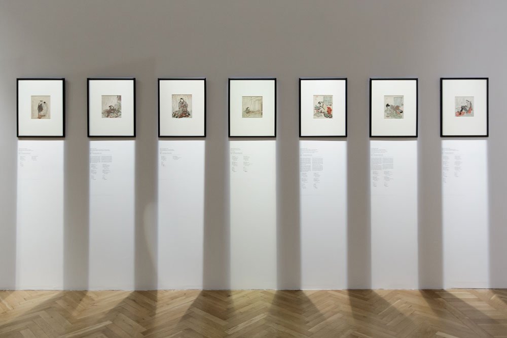 Siedem oprawionych grafik w czarnych ramkach równomiernie rozmieszczonych na jasnoszarej ścianie galerii, pod każdą ramką oświetlony biały panel z tekstem informacyjnym i widocznymi pionowymi cieniami, na dole jasna podłoga z drewnianego parkietu ułożonego w jodełkę.