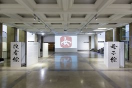 Duża, jasna sala wystawowa z kasetonowym sufitem i błyszczącą posadzką, centralnie ekran z czerwonym logo odbijającym się na podłodze, po obu stronach stoją pionowe panele z dużymi znakami kaligraficznymi oraz sufitowe lampy i kolumny.