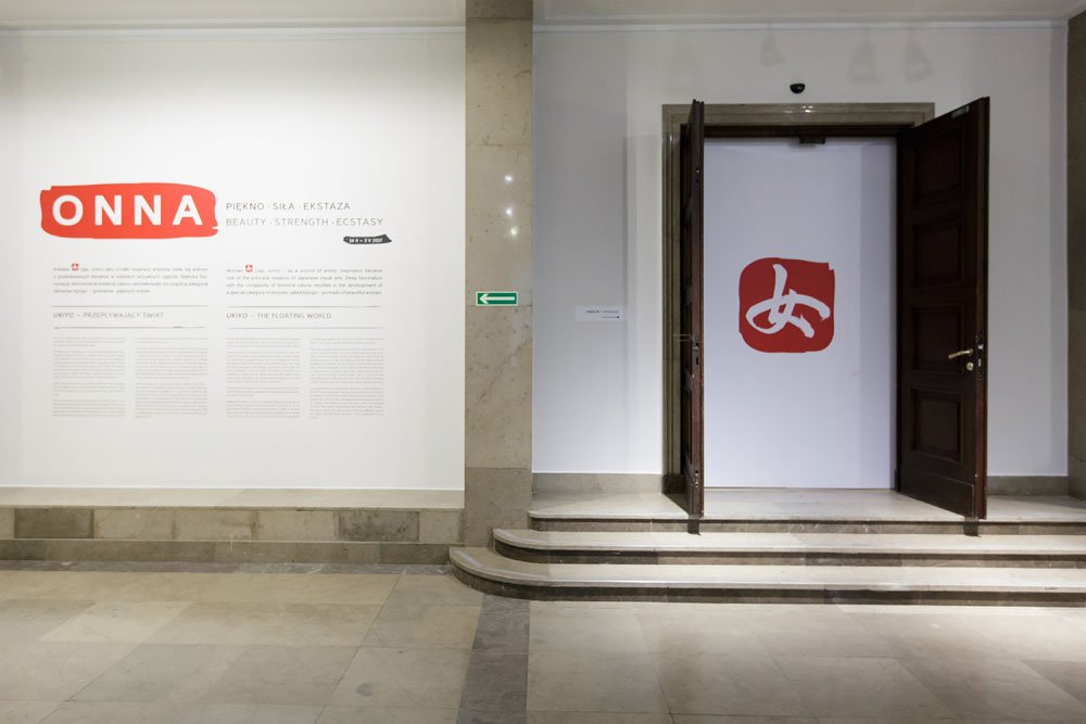 Wnętrze galerii z białą ścianą po lewej z dużym czerwonym napisem ONNA i tekstem informacyjnym, środkową kamienną kolumną z zieloną strzałką ewakuacyjną, po prawej kilkoma stopniami i otwartymi ciemnymi drewnianymi drzwiami prowadzącymi do jasnej sali z czerwonym znakiem na białej ścianie oraz beżową kamienną podłogą.
