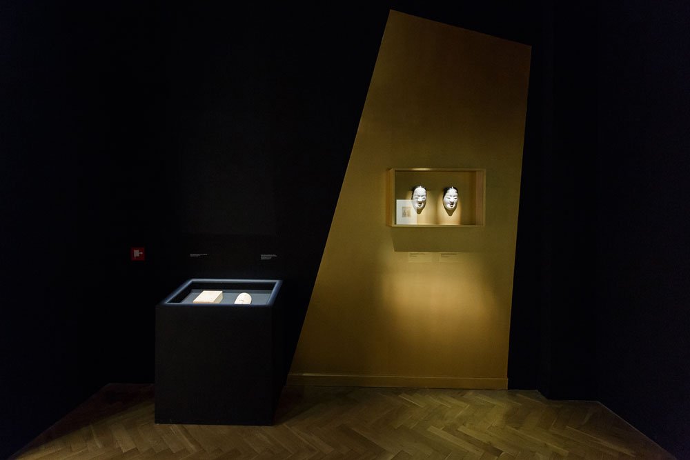 Ciemne wnętrze galerii z drewnianą chevronową podłogą, ukośnie ustawioną złotą ścianką po prawej z podświetloną wnęką eksponującą dwie białe maski oraz po lewej niską czarną przeszkloną witryną z podświetlonym dokumentem i małym obiektem.