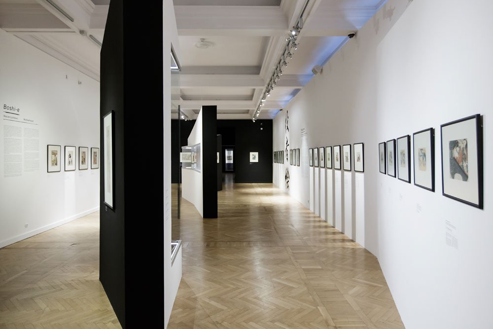 Wnętrze nowoczesnej galerii z jasnym drewnianym parkietem, białymi i czarnymi ścianami oraz wolnostojącymi czarnymi panelami, po prawej długim rzędem oprawionych w ramki prac wiszących i oświetleniem sufitowym.