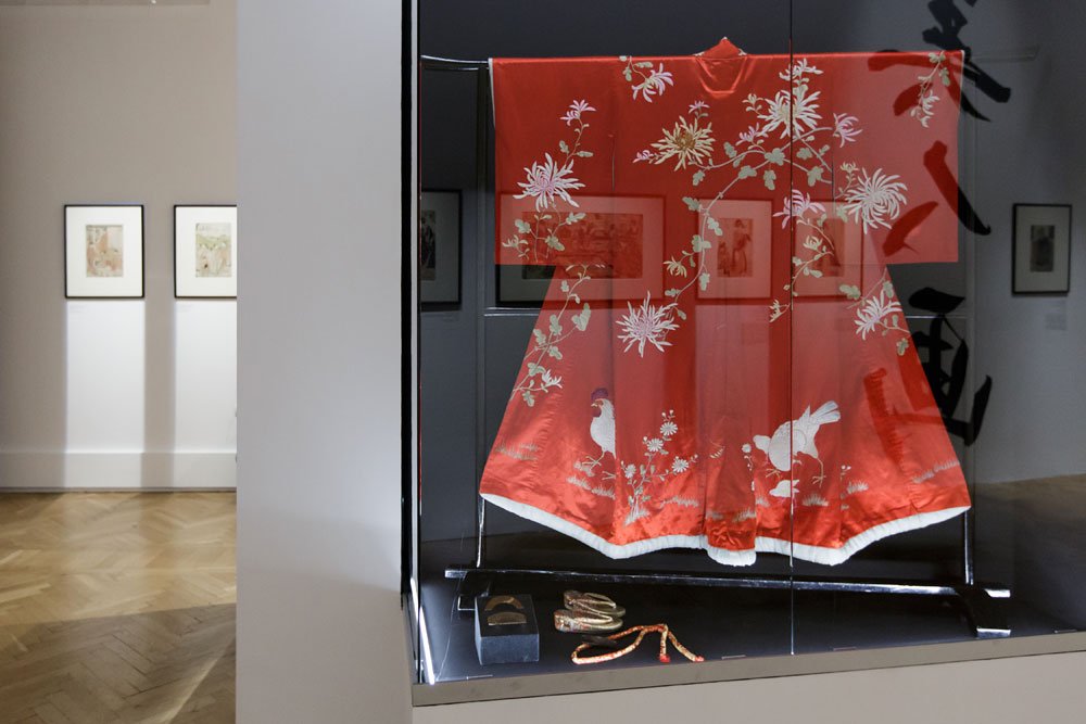 Czerwone haftowane kimono z motywami kwiatów i ptaków zawieszone za szkłem w muzealnej gablocie, z drobnymi akcesoriami (buty i sznurki) na półce poniżej oraz widoczną po lewej stronach częścią galerii z oprawionymi rycinami i parkietową podłogą.