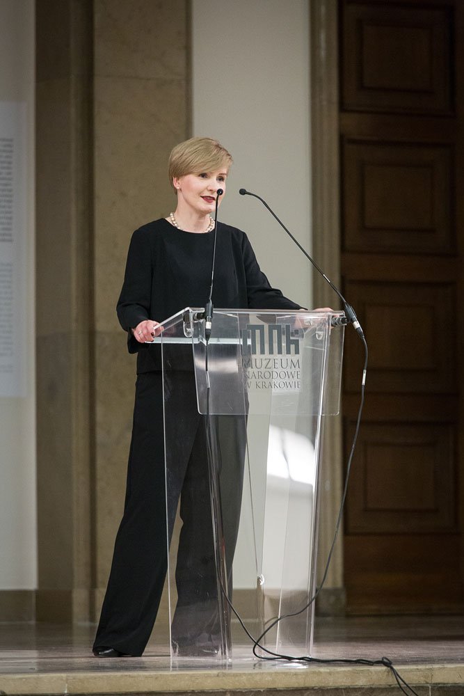 Kobieta o krótkich blond włosach w czarnym stroju i naszyjniku stoi za przezroczystą mównicą z napisem "Muzeum Narodowe w Krakowie" i mówi do mikrofonów w sali z kamiennym filarem i drewnianymi drzwiami, na podłodze widoczne kable.