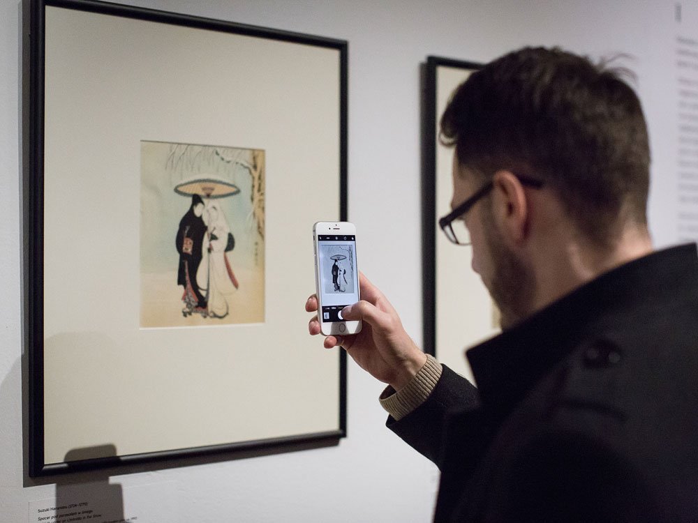 Mężczyzna widoczny z profilu trzyma smartfon i fotografuje oprawioną grafikę na ścianie galerii — w ramie z jasnym passe-partout znajduje się ilustracja przedstawiająca dwie postacie pod dużym parasolem, a na ekranie telefonu widać podgląd tej samej grafiki; mężczyzna ma krótkie włosy, okulary, ciemny płaszcz i krótką brodę.
