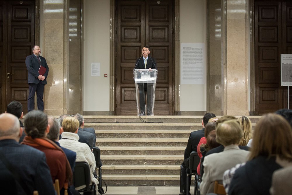 Mężczyzna w garniturze przemawia przy przezroczystej mównicy ustawionej na marmurowych schodach przed dużymi drewnianymi drzwiami w reprezentacyjnym holu, po lewej stoi inny mężczyzna z teczką, a na pierwszym planie widoczne są rzędy siedzącej publiczności zwróconej ku mówcy.