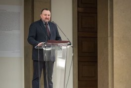 Mężczyzna w ciemnym garniturze i krawacie stoi za przezroczystą mównicą z mikrofonem i dokumentami w jasnym wnętrzu, w tle drewniane drzwi i kamienna kolumna.