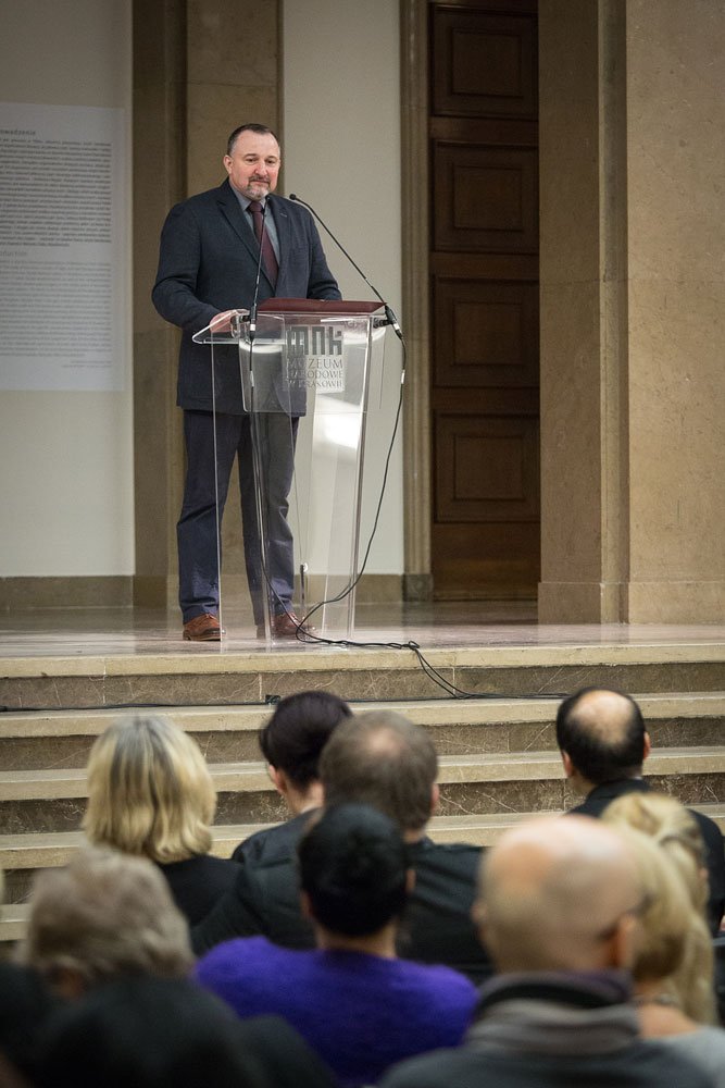 Mężczyzna w ciemnym garniturze i krawacie stoi za przezroczystą mównicą z mikrofonami na podwyższeniu sali z marmurowymi stopniami i drewnianymi drzwiami w tle, a z przodu widać część publiczności siedzącej tyłem.