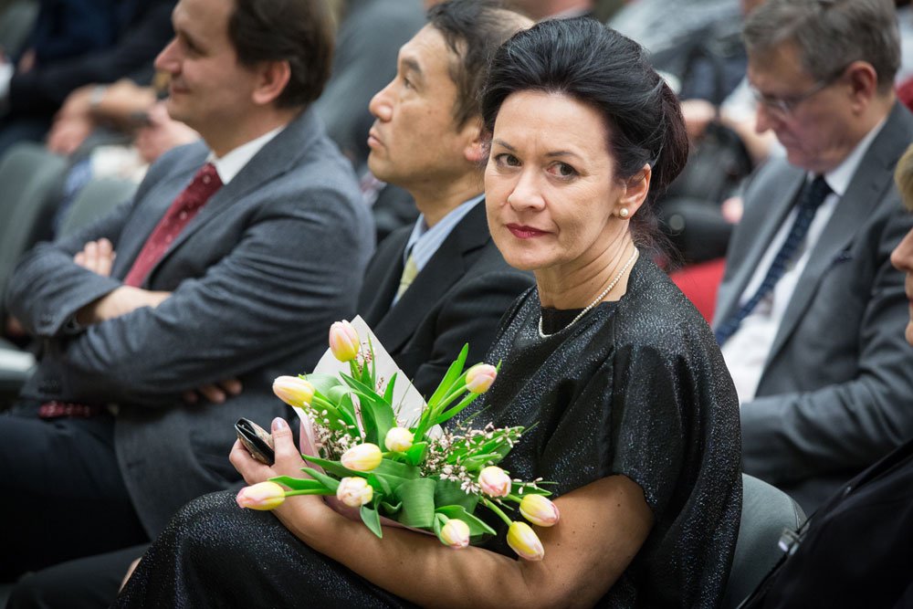 Kobieta w ciemnej połyskującej sukni i perłowych kolczykach siedzi wśród innych osób w sali, trzyma bukiet różowo‑żółtych tulipanów i telefon, patrząc w kierunku aparatu, a w tle widać kilku mężczyzn w garniturach.