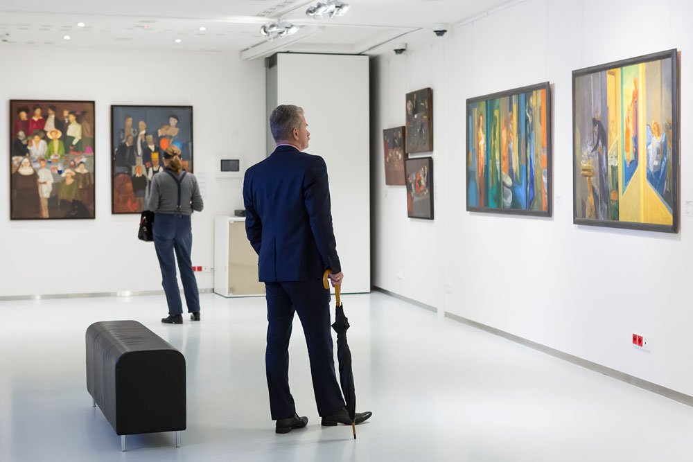 Mężczyzna w granatowym garniturze trzymający złożony czarny parasol stoi w jasnej galerii sztuki i ogląda obrazy na białych ścianach; w tle druga osoba przy dwóch dużych obrazach po lewej oraz niska czarna ławka na środku pomieszczenia.