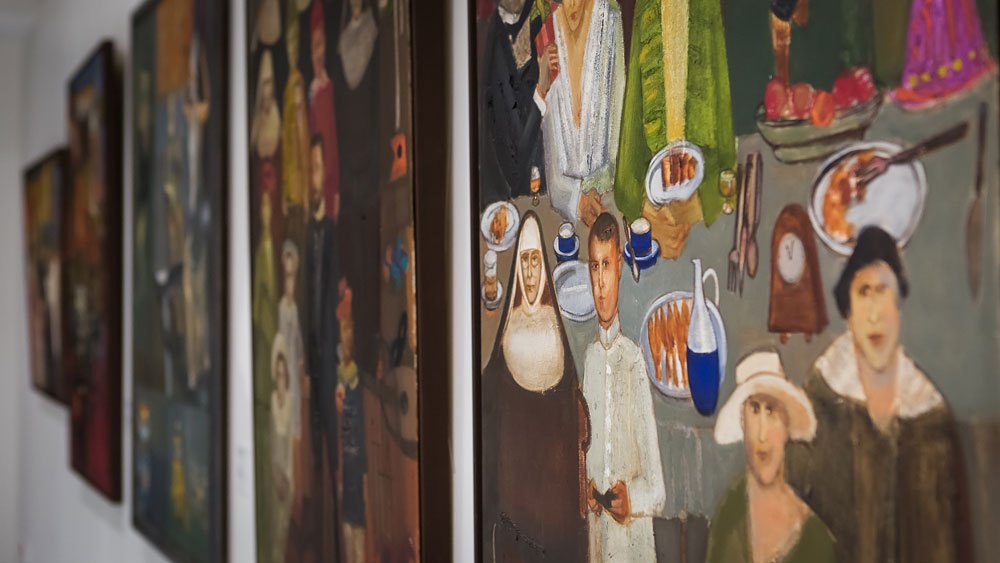 Zbliżenie na kilka obrazów wiszących w galerii; na pierwszym planie kolorowy, figuratywny obraz przedstawiający ludzi siedzących przy stole z naczyniami i jedzeniem — m.in. postać ubrana w habit, chłopiec w białej koszuli, dzban i talerze — a w tle rozmyte ramy kolejnych obrazów na białej ścianie.