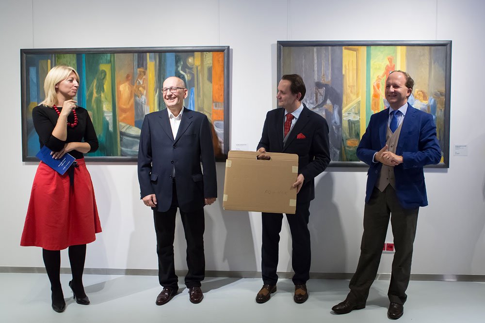 W galerii na białej ścianie wiszą dwa kolorowe, ramowane obrazy, a przed nimi stoją cztery osoby: po lewej kobieta w czarnej bluzce i czerwonej spódnicy z czerwonym naszyjnikiem trzymająca niebieską książeczkę, obok starszy mężczyzna w ciemnym garniturze uśmiechający się, dalej mężczyzna w ciemnym garniturze i czerwonym krawacie trzymający kartonowe pudełko, a po prawej mężczyzna w niebieskiej marynarce i beżowej kamizelce.