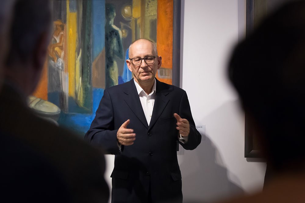 Mężczyzna w ciemnym garniturze i okularach stoi przed kolorowym, abstrakcyjno‑figuracyjnym obrazem w galerii i gestykuluje rękami, na pierwszym planie rozmyte sylwetki widzów.