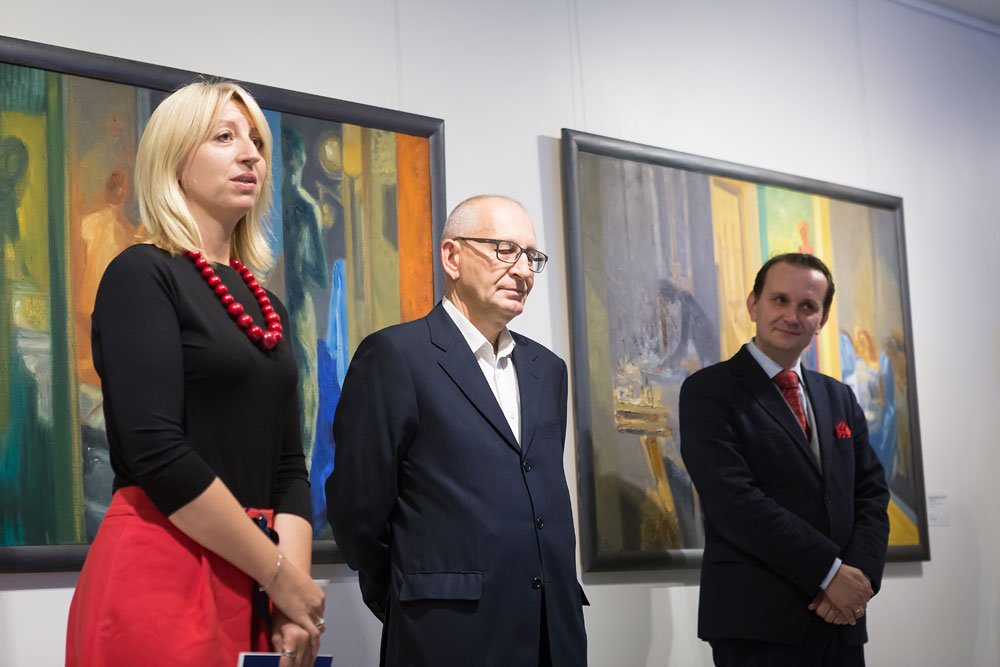 Troje osób stoi obok siebie przed dwoma dużymi oprawionymi obrazami na białej ścianie galerii: z lewej blondynka w czarnej bluzce, czerwonej spódnicy i naszyjniku z dużych czerwonych korali trzymająca niebieską kartkę, w środku starszy mężczyzna w ciemnym garniturze i okularach z rękami za plecami, z prawej mężczyzna w ciemnym garniturze z czerwonym krawatem i poszetką, z rękami złożonymi.