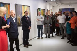 Wnętrze jasnej galerii z kilkoma kolorowymi, oprawionymi obrazami na ścianach; grupa osób stoi wzdłuż ścian, po lewej blond kobieta trzyma mikrofon, obok niej starszy mężczyzna i mężczyzna w ciemnym garniturze, po prawej rząd ludzi w różnorodnych strojach.