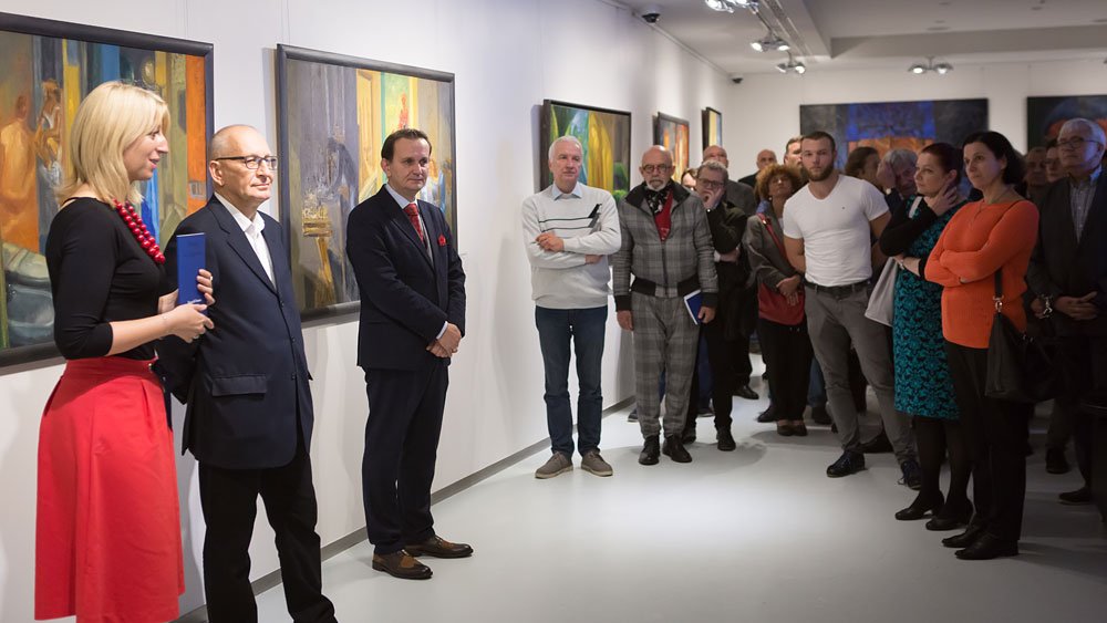 Wnętrze galerii sztuki z białymi ścianami i oprawionymi obrazami, na pierwszym planie po lewej kobieta w czerwonej spódnicy i czarnej bluzce trzymająca niebieski przedmiot, obok dwóch mężczyzn w garniturach, a po prawej zgromadzona grupa widzów w różnym wieku obserwująca wystawę.
