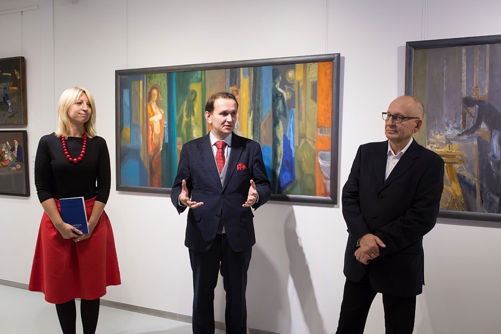Troje osób stoi w białej galerii przed oprawionymi obrazami: po lewej kobieta w czarnej bluzce i czerwonej spódnicy trzyma niebieską książkę i nosi czerwony naszyjnik, w środku mężczyzna w granatowym garniturze z czerwonym krawatem gestykuluje, po prawej mężczyzna w ciemnym garniturze stoi z założonymi rękami, a za nimi wisi duży kolorowy obraz figuralny.