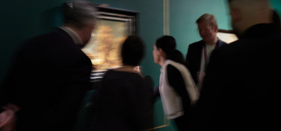 Kilka rozmytych osób w ciemnych ubraniach, jedna w białej kamizelce, pochylonych nad małym oprawionym obrazem zawieszonym na zielonej ścianie.