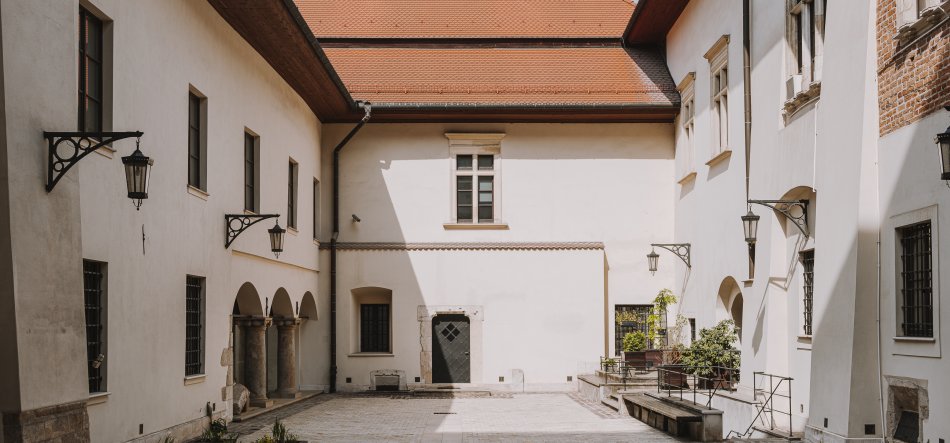Pusty kamienny dziedziniec otoczony jasnymi elewacjami budynku z czerwoną dachówką, po lewej łukowe przejścia z kolumnami i metalowe kinkiety, po prawej ławki i donice z roślinami, liczne okna z kratami oraz centralne drzwi z widocznym wzorem i cieniem padającym na ścianę.