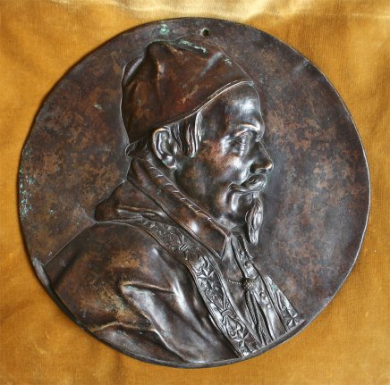 Okrągła brązowa płaskorzeźba przedstawiająca popiersie mężczyzny w profilu zwróconego w prawo, z czapką, wąsami i brodą oraz ozdobnym strojem z reliefowanym wzorem, z widoczną patyną i niewielkim otworem u górnej krawędzi, umieszczona na złotym aksamitnym tle.