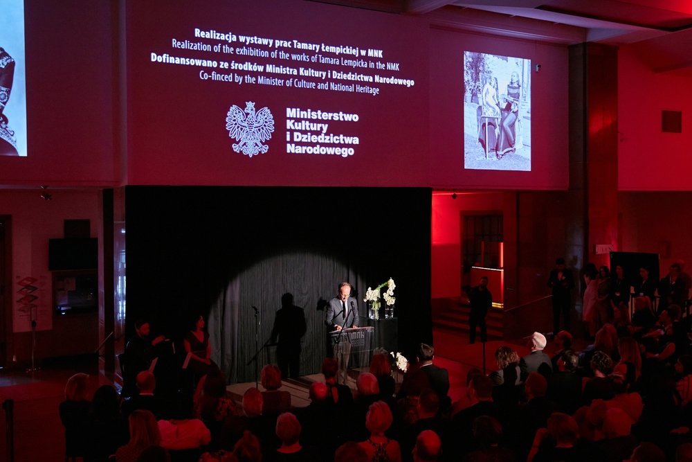 Sala z publicznością o czerwonym oświetleniu, męski mówca stoi przy mównicy na niewielkiej scenie ozdobionej kwiatami, nad sceną duży ekran z napisem "Ministerstwo Kultury i Dziedzictwa Narodowego" i czarno-białymi zdjęciami, widok z tyłu na zebranych.