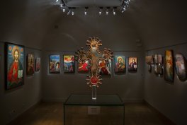 Niewielka, przyciemniona sala wystawowa o półkolistym sklepieniu z punktowym oświetleniem, na ścianach ułożone kolorowe ikony w ramach, a na środku na szklanym postumencie wyeksponowany ozdobny krucyfiks.