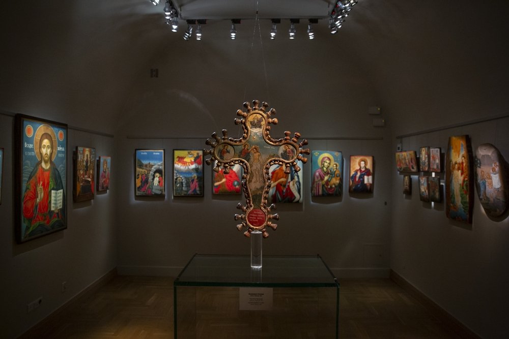 Widok wnętrza galerii z centralnie zawieszoną ozdobną, malowaną relikwią w kształcie krzyża nad szklanym postumentem, otoczoną na ścianach kolorowymi ikonami i religijnymi malowidłami oświetlonymi reflektorami.