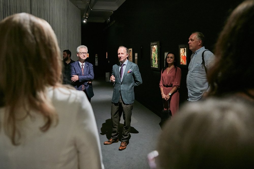 Grupa osób w ciemnej galerii sztuki: mężczyzna w szarej marynarce stoi na środku z lekko uniesioną dłonią, obok niego stoi starszy mężczyzna w garniturze z założonymi rękami, po prawej kobieta w różowej sukience i mężczyzna w jasnej koszuli, z przodu widać rozmyte sylwetki widzów, a na czarnej ścianie za nimi wiszą oświetlone obrazy w ramach.