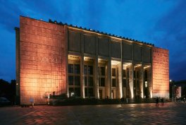 Monumentalny kamienny budynek o podświetlonej na ciepło fasadzie z kilkoma wysokimi pionowymi kolumnami i dużymi oknami, stojący przy pustym, szerokim placu pod głębokim niebem o zmierzchu.