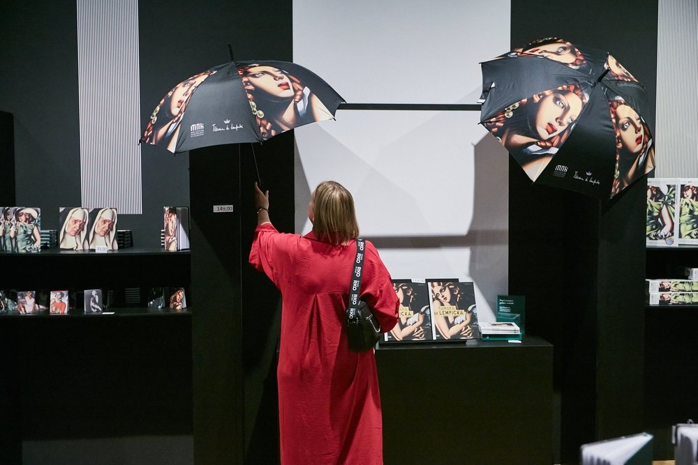Osoba widziana od tyłu w długiej czerwonej sukience podnosi wiszący czarny parasol z dużym nadrukiem twarzy kobiety; obok wisi drugi podobny parasol nad czarnymi półkami z książkami i gadżetami z tym samym motywem, na których widoczna jest metka z ceną 149,00.