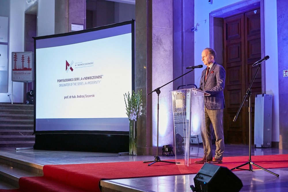 Mężczyzna stojący przy przezroczystym pulpicie z mikrofonem na podwyższeniu obok dużego ekranu projekcyjnego po lewej z napisem o serii „4 x nowoczesność”, czerwony dywan na scenie, wazon z białymi kwiatami oraz schody i drewniane drzwi w tle.