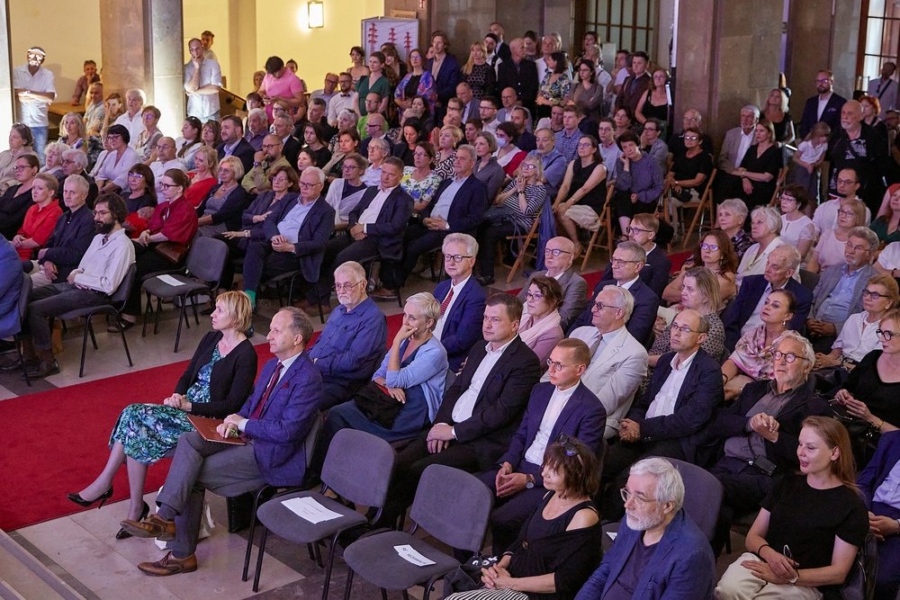 Duża grupa osób w różnym wieku siedzi w rzędach krzeseł i stoi z tyłu w dużej sali z kamiennymi kolumnami i czerwonym dywanem przy lewej krawędzi, większość ubrana półformalnie i zwrócona w stronę przodu.