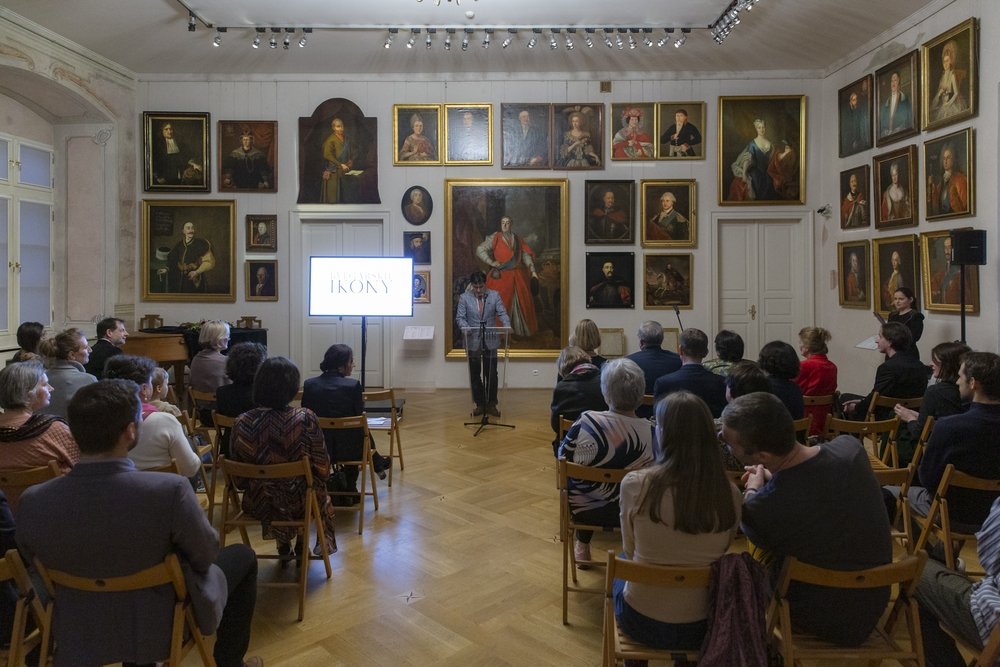 Sala z drewnianą podłogą i ścianami wypełnionymi licznymi portretami w złotych ramach, w której osoba stoi przy mównicy obok ekranu prezentacyjnego, a przed nią rzędy publiczności siedzącej na składanych krzesłach.