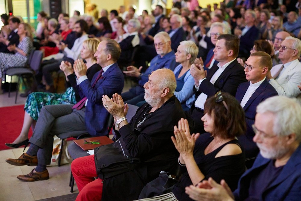Publiczność siedząca w rzędach w sali — w pierwszym planie starszy mężczyzna z siwą brodą w czarnej kurtce i czerwonych spodniach oraz kobieta w czarnym topie; wszyscy widoczni uczestnicy biją brawa, w tle kolejne rzędy osób w formalnych ubraniach.