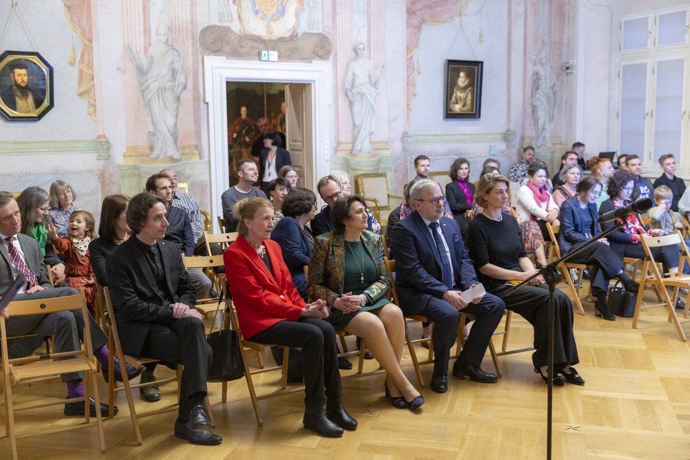 Grupa osób siedzi na składanych drewnianych krzesłach w zdobnej sali z malowanymi freskami i obrazami na ścianach, na pierwszym planie rząd osób — mężczyzna w czarnym stroju, kobieta w czerwonej marynarce, kobieta w zielonej sukience z wzorzystą marynarką, starszy mężczyzna w garniturze i kobieta w czarnej sukience — w tle kolejne rzędy widzów, otwarte drzwi z przechodzącymi osobami oraz stojący z przodu mikrofon.