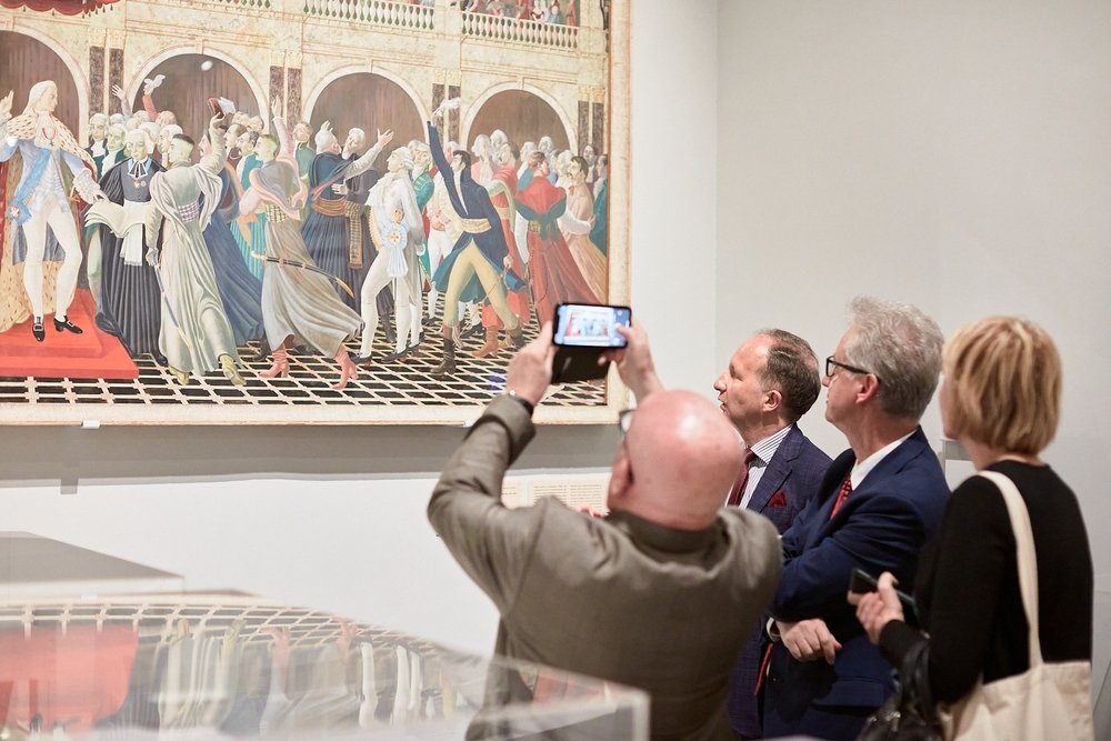 Na zdjęciu w galerii cztery osoby stoją przed dużym, ramowanym obrazem historycznym na białej ścianie; mężczyzna na pierwszym planie robi zdjęcie smartfonem, u dołu widać odbicie w przeszklonej gablocie.