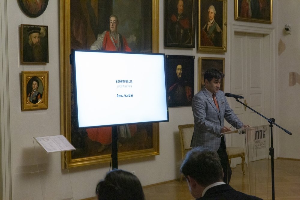 Mężczyzna w kratowanej marynarce przemawia przy przezroczystej mównicy z mikrofonem, obok stoi duży ekran na statywie wyświetlający biały slajd z napisem "Koordynacja" i "Anna Gardaś", a w tle ściana udekorowana wieloma portretami w ozdobnych ramach; na pierwszym planie widoczne sylwetki słuchaczy.