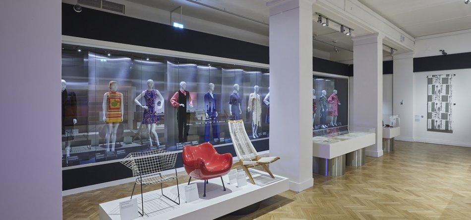 Wnętrze jasnej galerii muzealnej z drewnianym parkietem i białymi filarami; w tle długa przeszklona witryna z szeregiem manekinów w kolorowych sukienkach i strojach, na pierwszym planie niski biały podest z trzema krzesłami — metalowym ażurowym, czerwonym formowanym i wiklinowo-drewnianym — oraz przeszklone gabloty wystawowe i czarno-biały obraz na prawej ścianie.