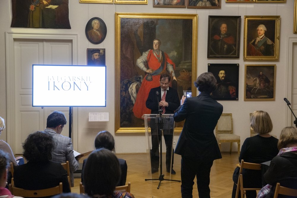 Mężczyzna przemawiający przy przezroczystym pulpicie przed dużym złoconym portretem w sali galerii z wieloma oprawionymi obrazami na ścianach, przed publicznością siedzącą na krzesłach, osoba z tyłu robiąca zdjęcie telefonem oraz po lewej jasny monitor z widocznym napisem.