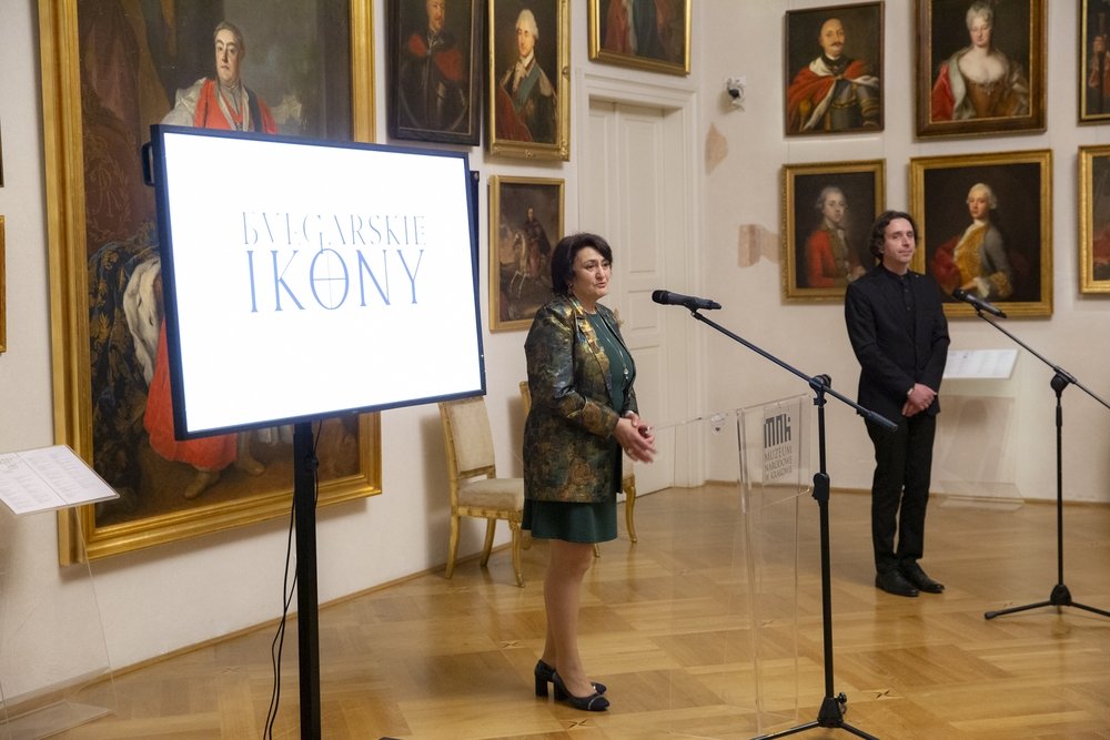 Wnętrze sali galerii z licznymi portretami w złotych ramach, po lewej duży ekran z napisem BUŁGARSKIE IKONY, na środku kobieta przy przezroczystym pulpicie z mikrofonem, po prawej stoi mężczyzna przy drugim mikrofonie.